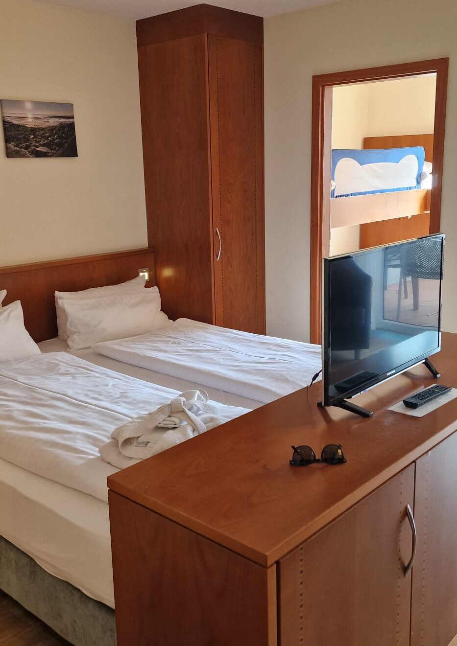 Doppelbett im Hotelzimmer, am Fußende steht eine Kommode, im hinteren Raum ein Doppelstockbett