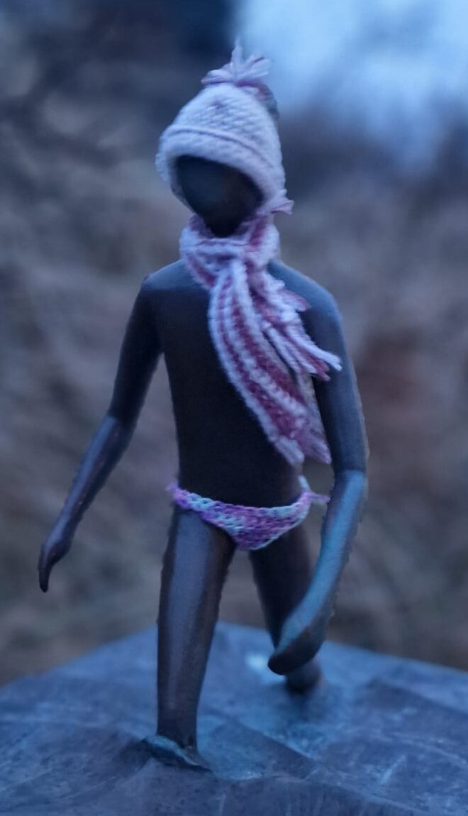 Bronzeskulptur einer Person die aus dem Meer läuft mit gehäkelter Badehose, Schal und Mütze
