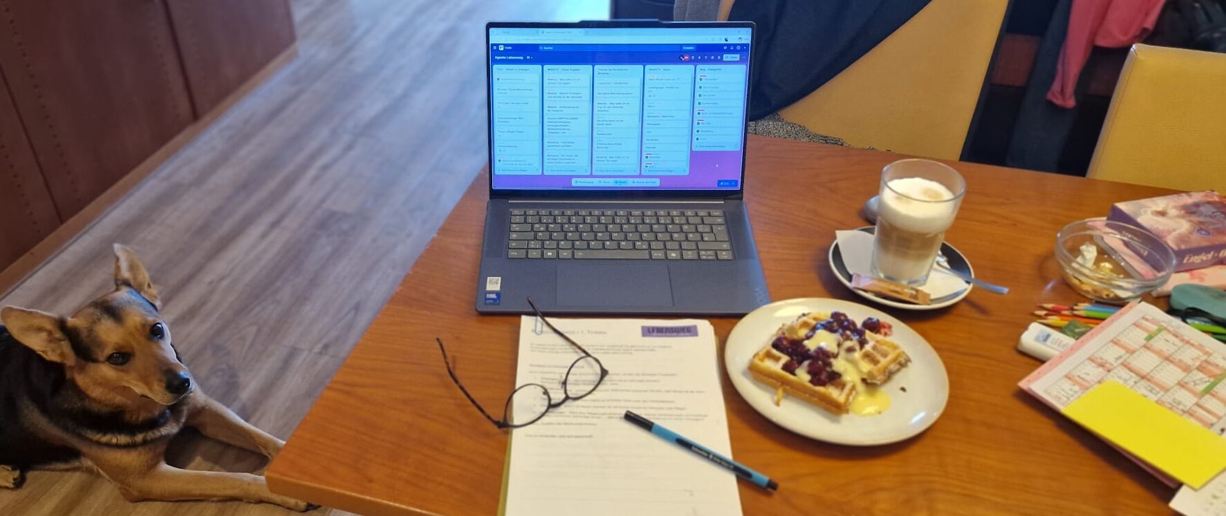 Laptop auf einem Tisch, Kaffee und ein Teller mit Waffel und Kirschen stehen daneben, am Boden liegt der Hund