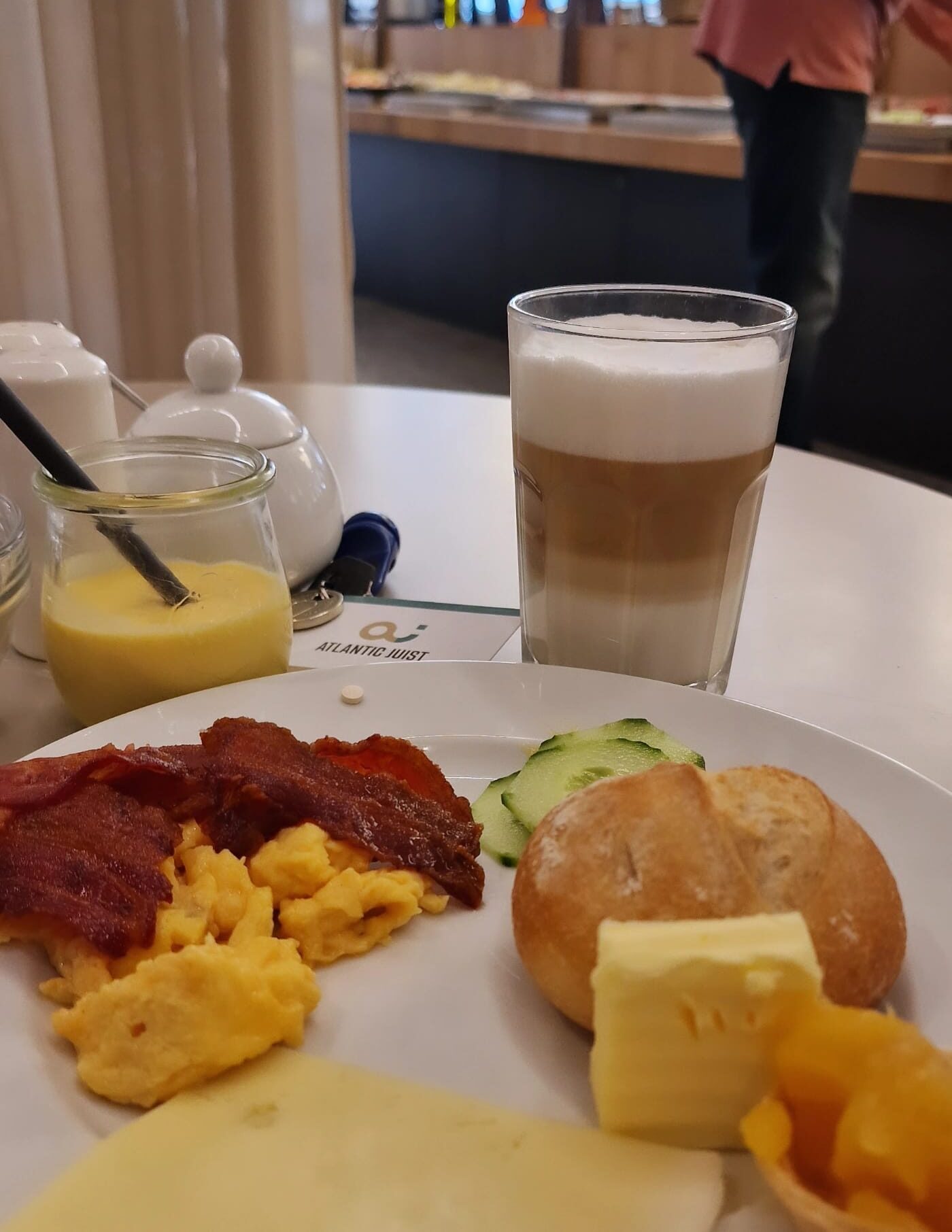 Teller mit Bacon, Rührei, Brötchen, Käse, Gurke, Butter, Marmelade, LatteMacchiato im Glas, Mango-Lassi im Glas mit Strohalm, Zuckerdose, Zimmerschlüssel, im Hintergrund das Buffet